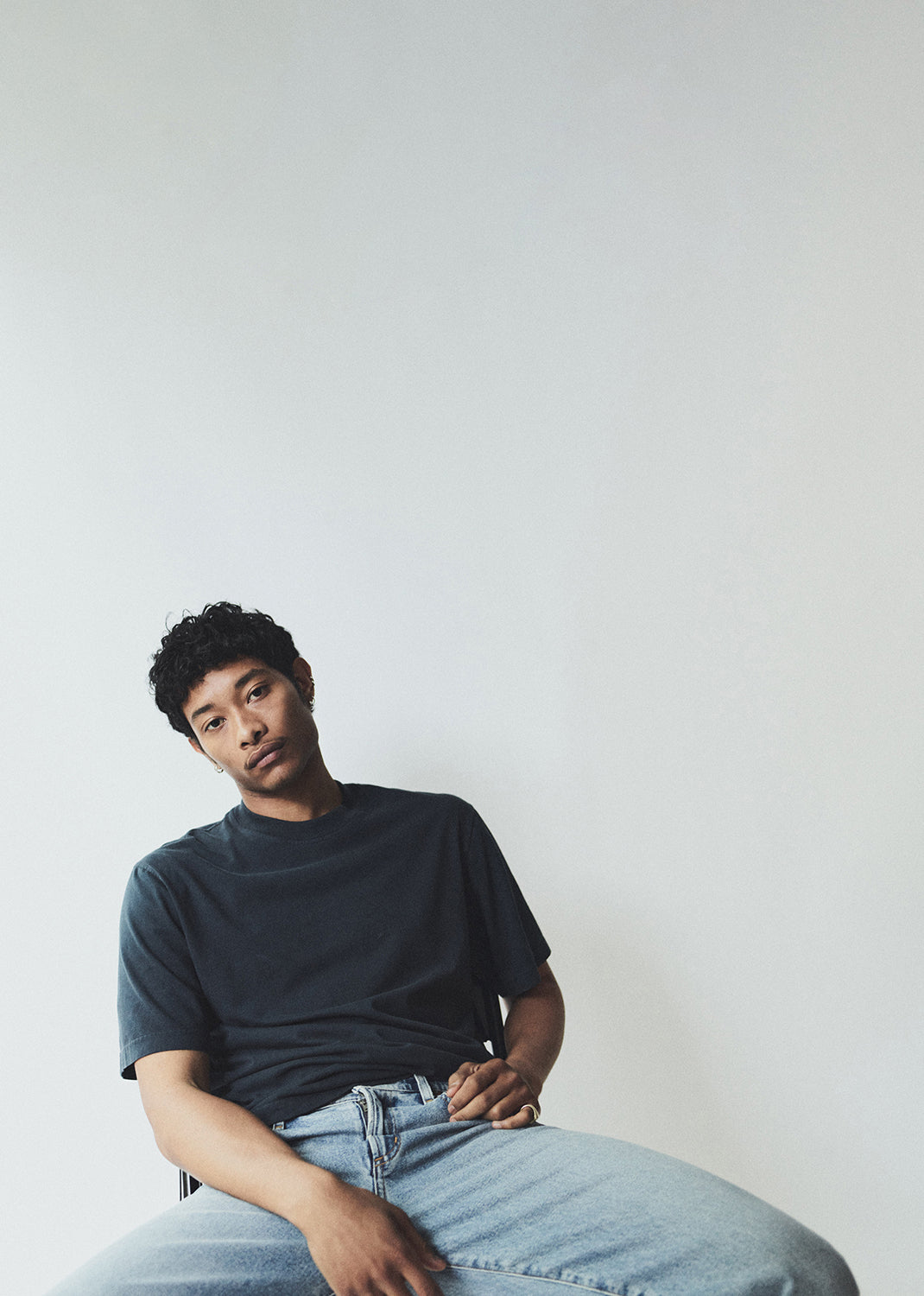 Person wearing a dark t-shirt and light jeans sitting against a plain background