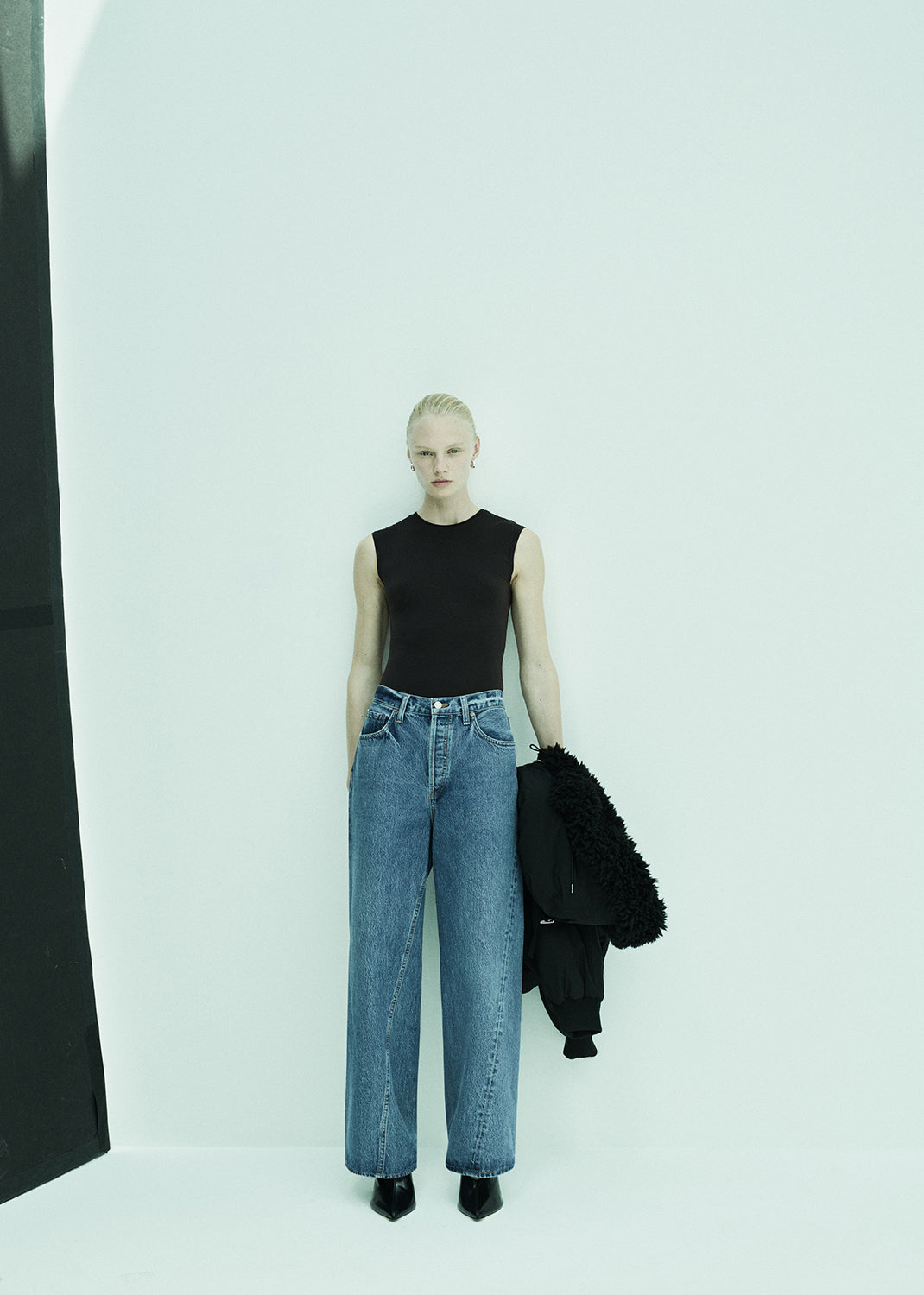 Person wearing a black sleeveless top and blue jeans holding a black jacket against a white background