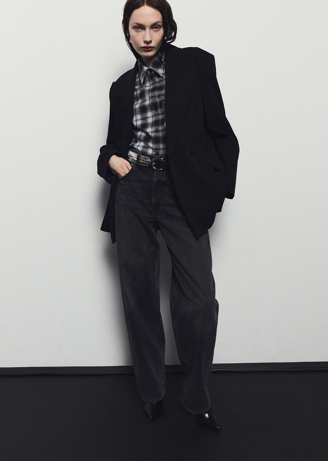 Person wearing a black coat and pants with a plaid shirt against a white wall.