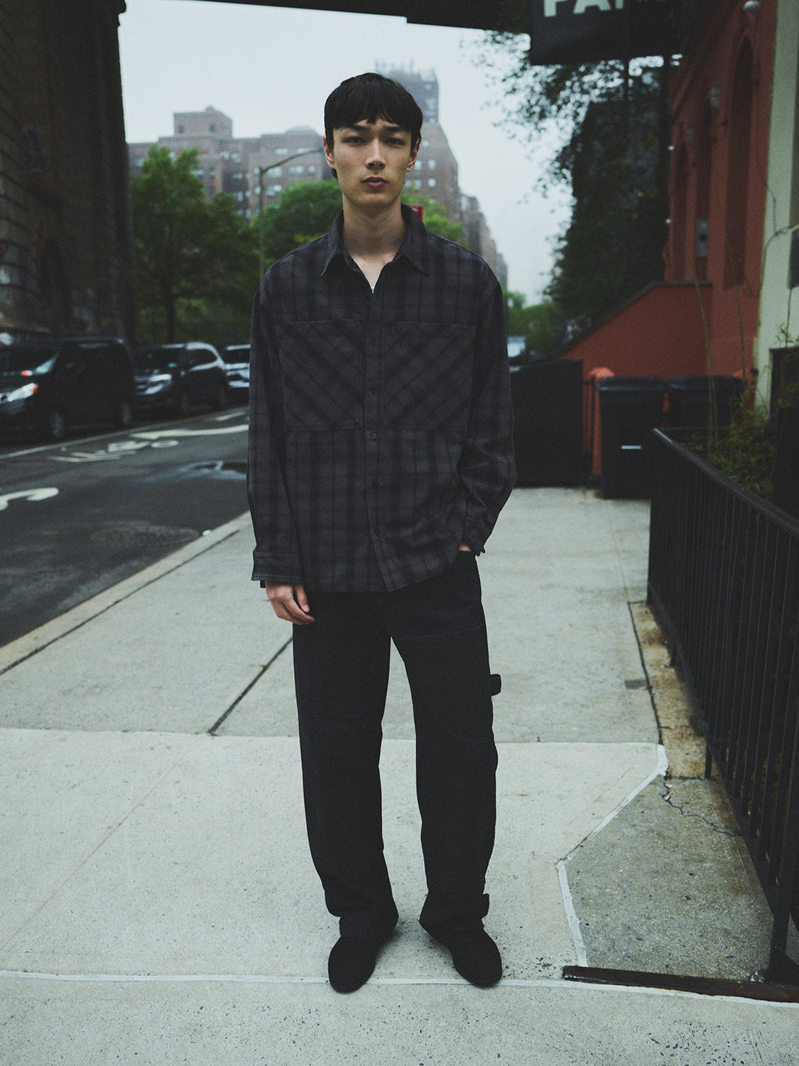 Person standing on a sidewalk wearing a dark plaid shirt and black pants.