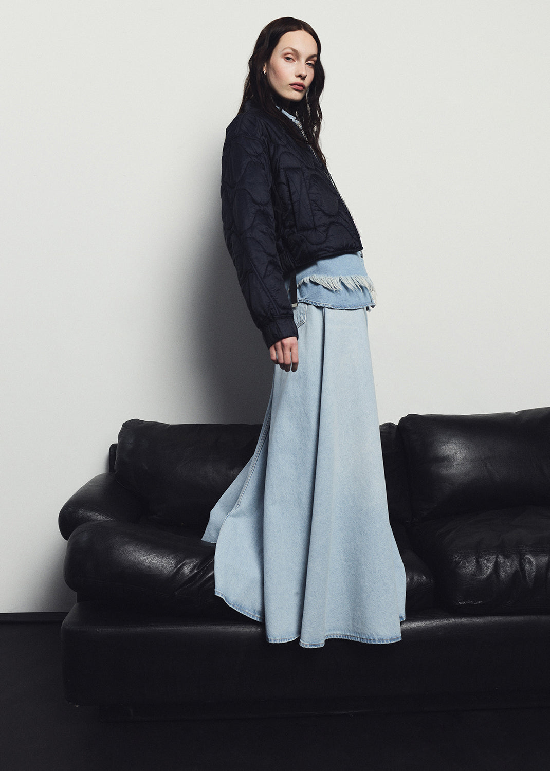 Woman in a black jacket and light blue denim skirt standing in front of a black leather couch against a white wall.