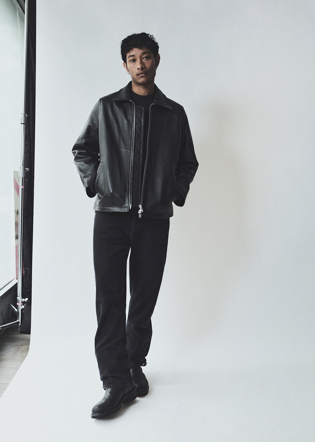 Man wearing a black leather jacket and pants standing against a white wall.