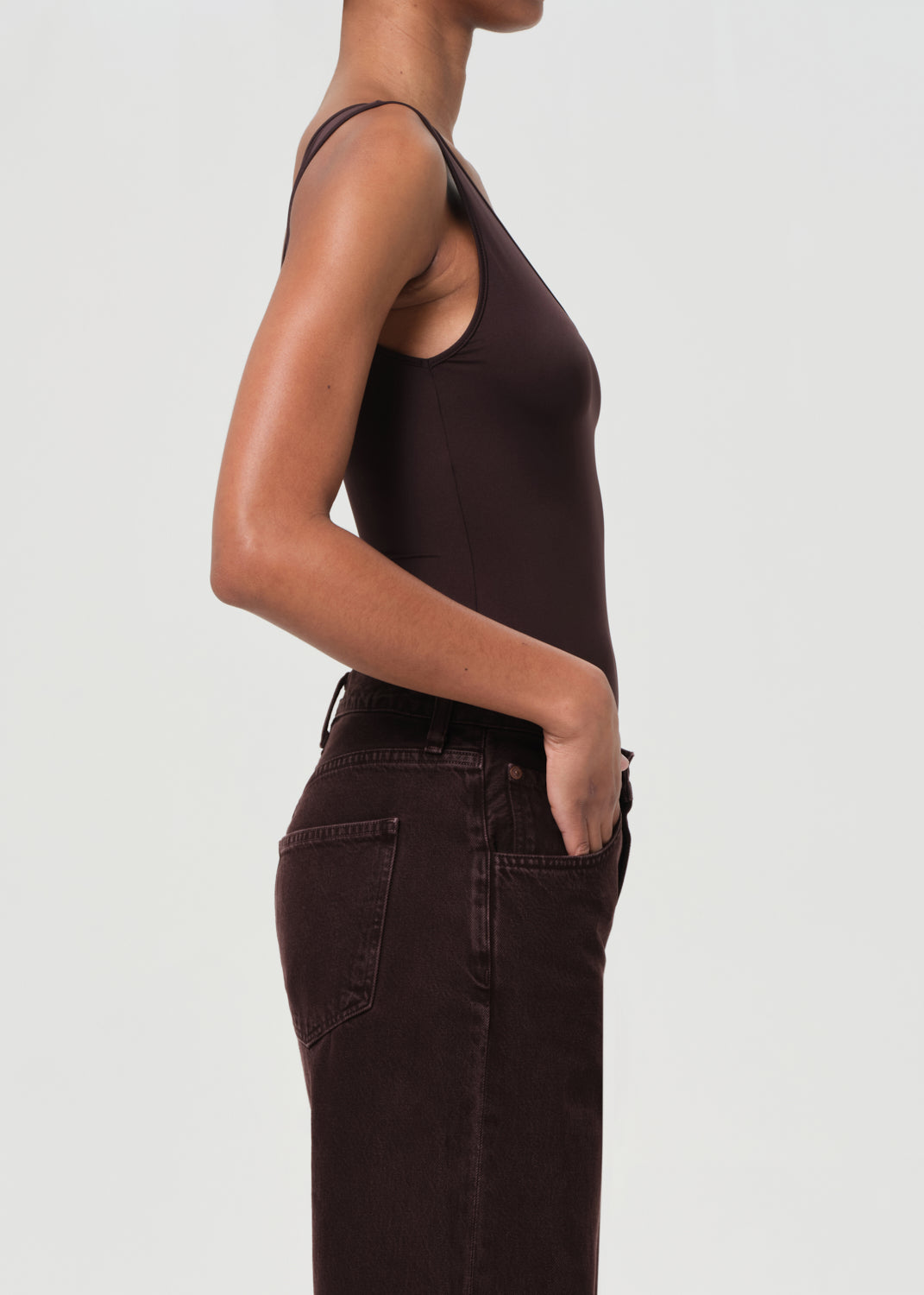 Person wearing a brown tank top and dark jeans on a light gray background