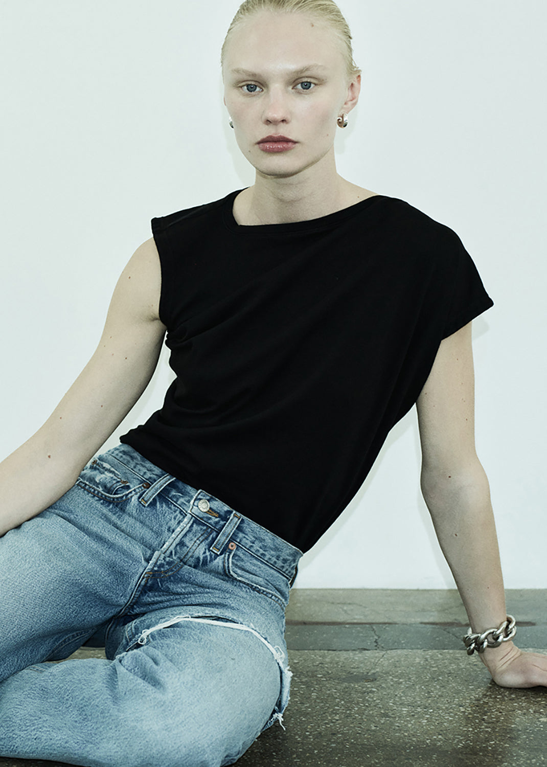 Person wearing a black sleeveless top and blue jeans sitting on a concrete surface.