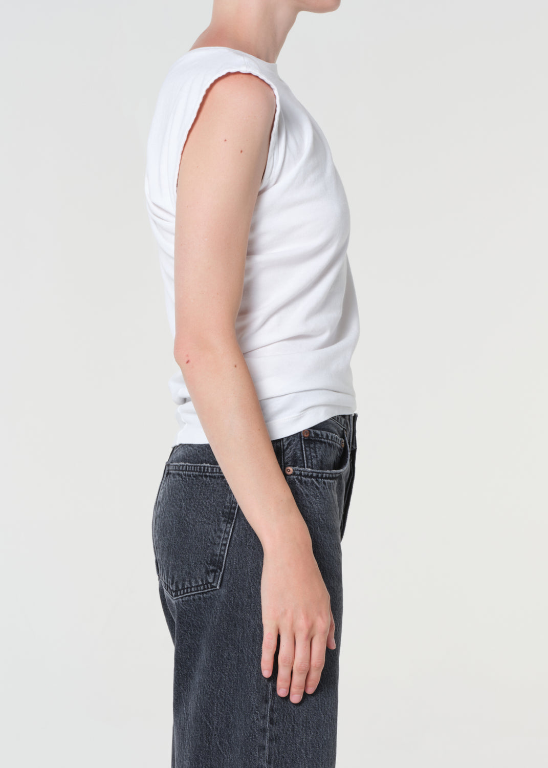 Person wearing a white sleeveless top and dark jeans on a plain background
