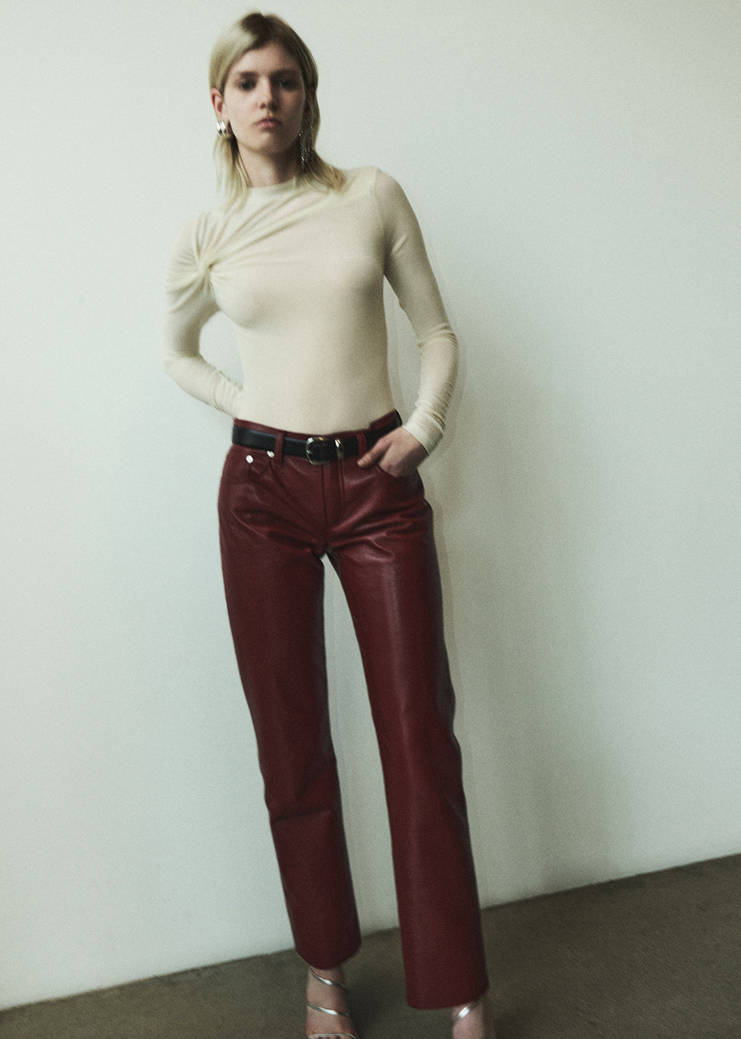 Woman wearing a beige long-sleeve top and red leather pants against a plain background