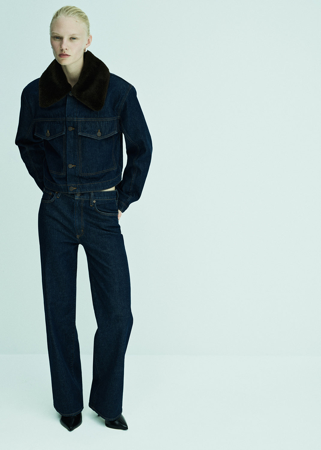 Person wearing a dark denim outfit with a fur-lined collar on a light blue background