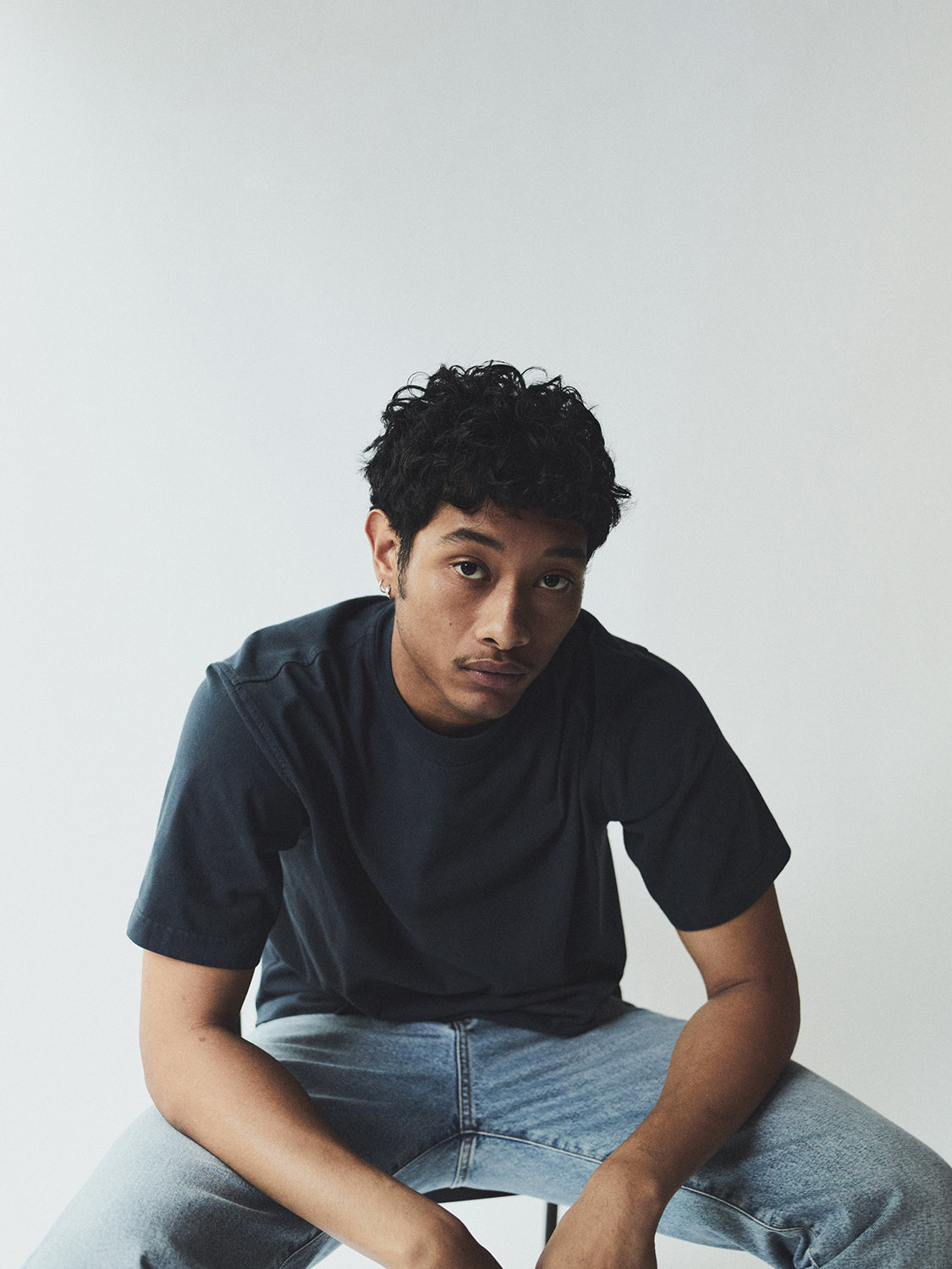 Person wearing a dark blue t-shirt  and light blue jeans sitting against a plain background