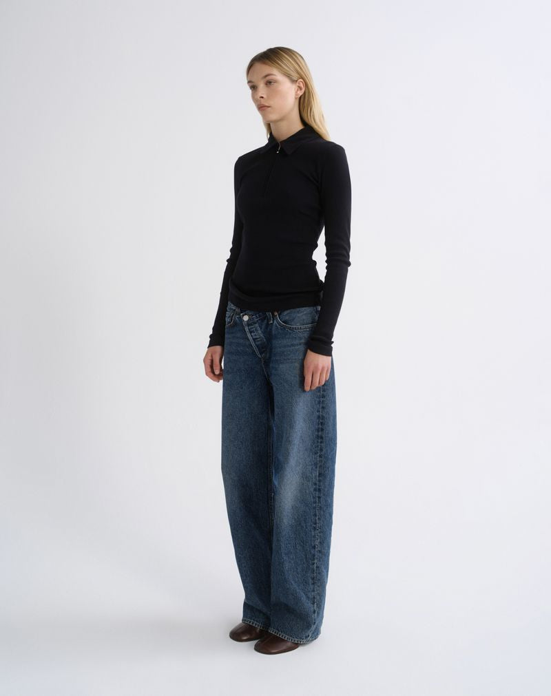 A woman modeling a black long-sleeve mock neck top and wide-leg blue jeans against a plain light gray studio background.