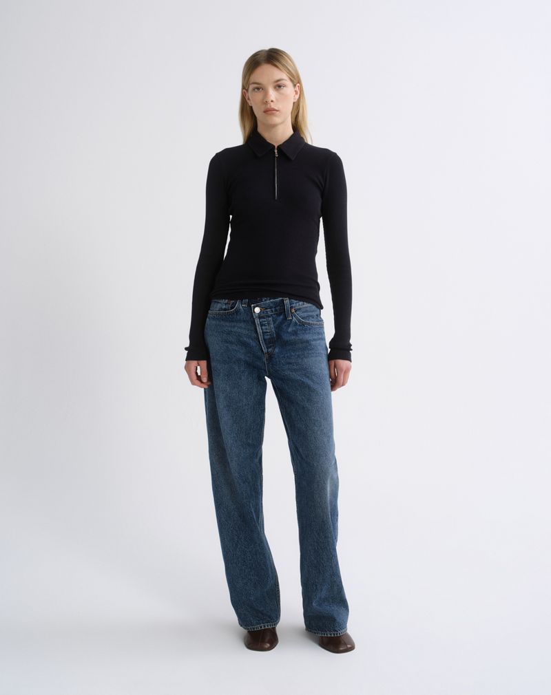 A woman stands against a light gray studio background wearing a fitted black long-sleeve zip-neck top, loose blue jeans, and dark brown shoes.