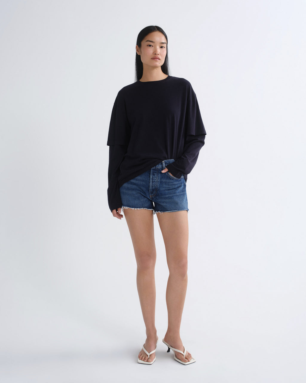 A woman modeling a black long-sleeve oversized top with blue denim shorts and white heeled sandals, standing against a plain light gray studio background.