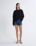 A woman modeling a black long-sleeve oversized top with blue denim shorts and white heeled sandals, standing against a plain light gray studio background.