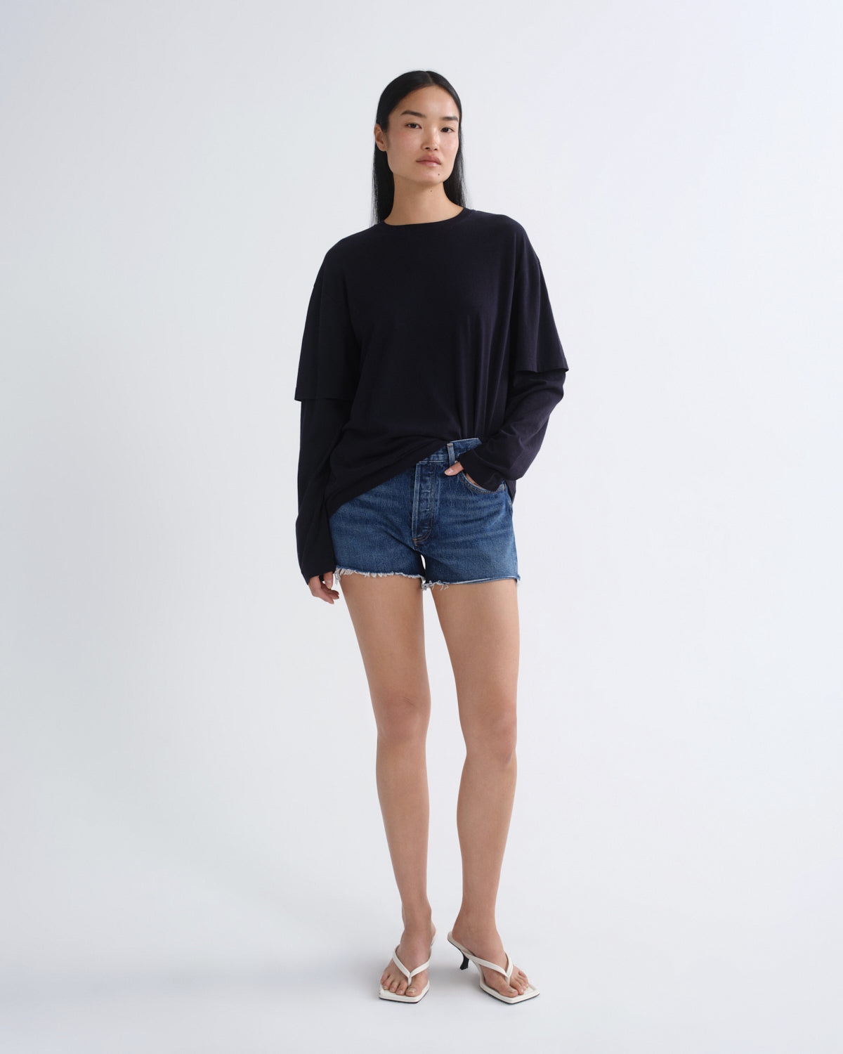 A woman modeling a black long-sleeve oversized top with blue denim shorts and white heeled sandals, standing against a plain light gray studio background.