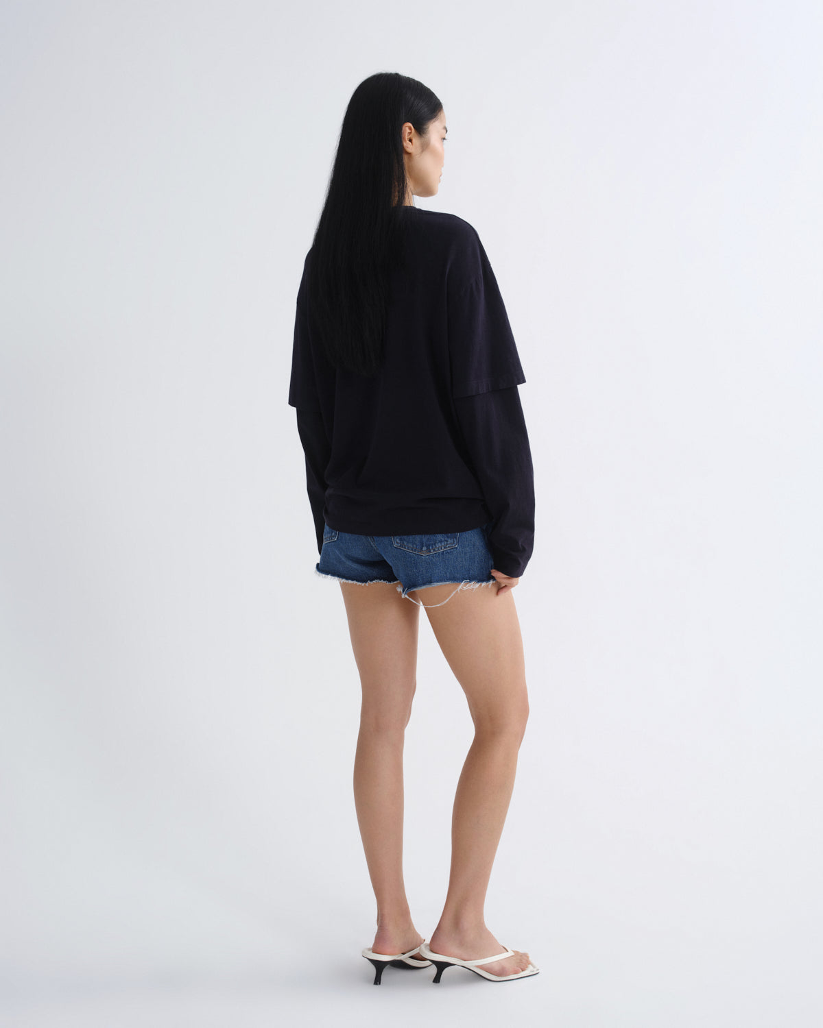 A woman is shown from the back wearing a black oversized long-sleeve top, blue denim shorts, and white heeled sandals against a plain light gray studio background.