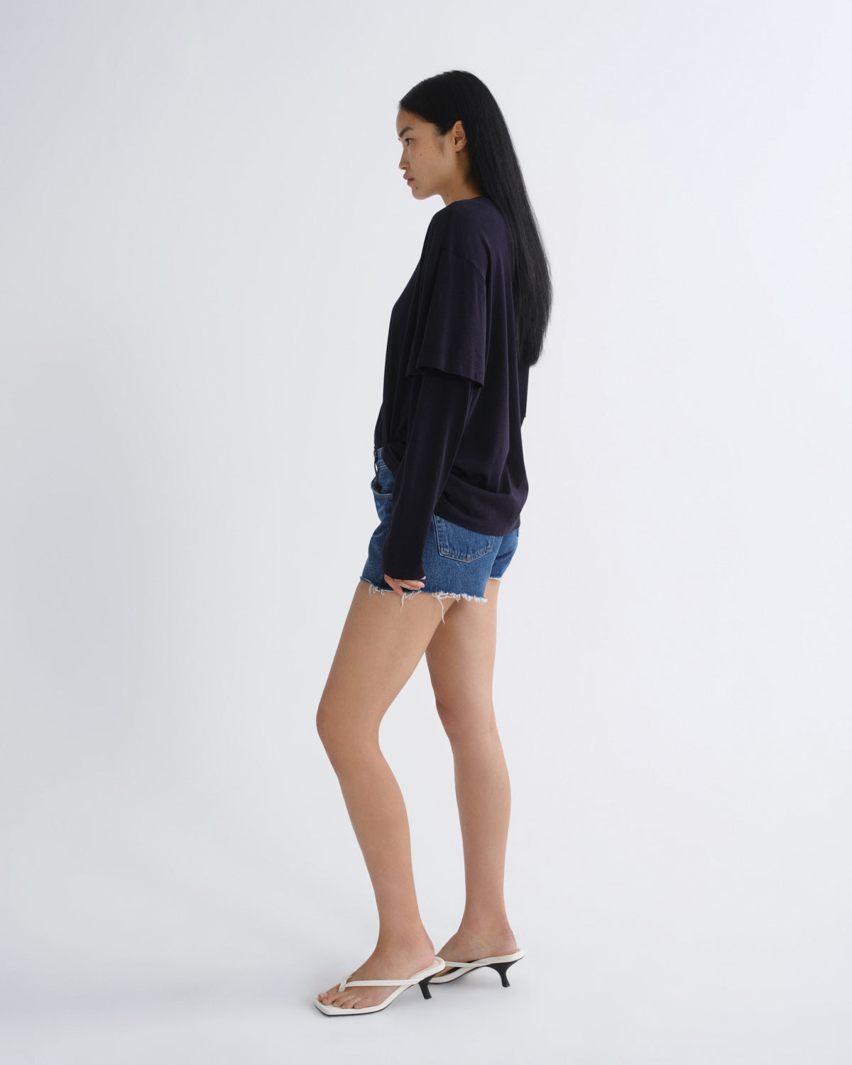 A woman stands in profile wearing a dark navy long-sleeve top, blue denim shorts, and white heeled thong sandals against a plain light gray studio background.