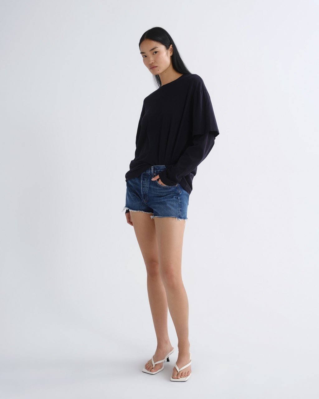 A model wearing a black long-sleeve top, blue denim shorts, and white heeled sandals, standing against a plain white studio background.
