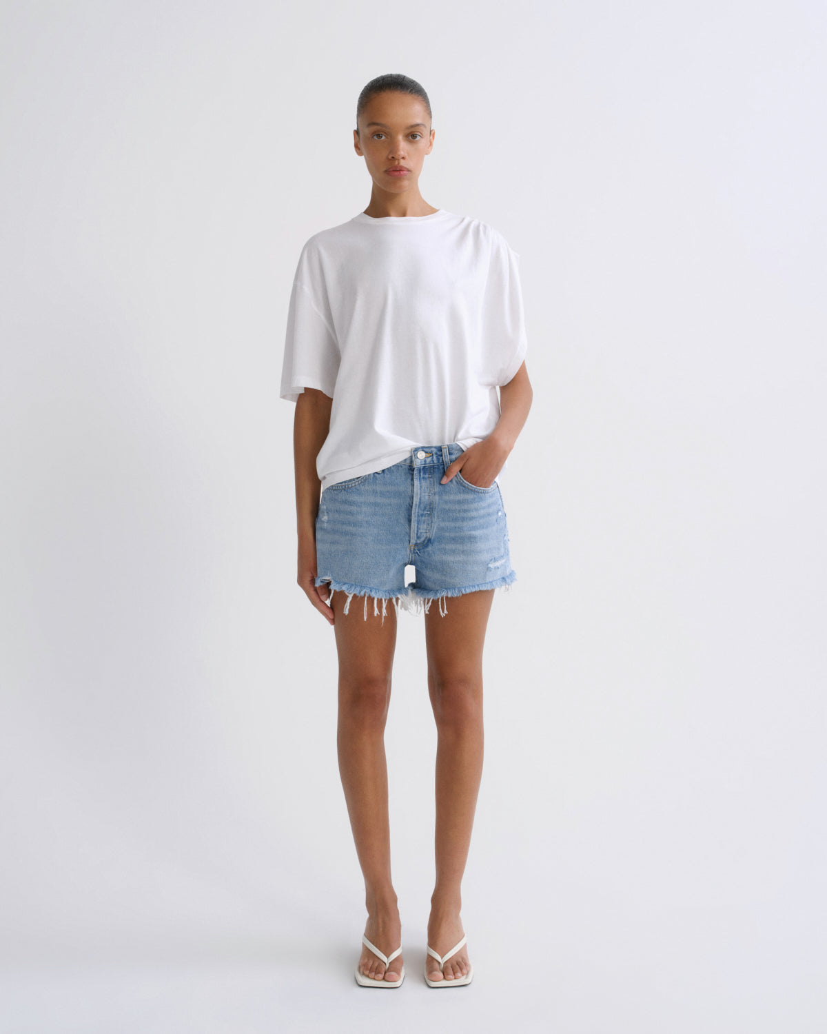 A model stands against a light gray studio background wearing a loose white short-sleeve t-shirt, light-wash blue denim shorts with frayed hems, and white thong sandals.