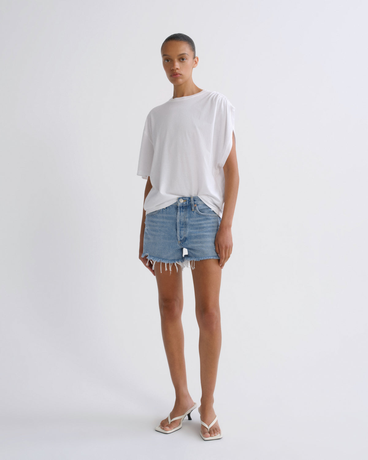 A model stands against a light gray studio background wearing a loose white oversized T-shirt, light-wash blue denim shorts with raw hems, and white heeled thong sandals.