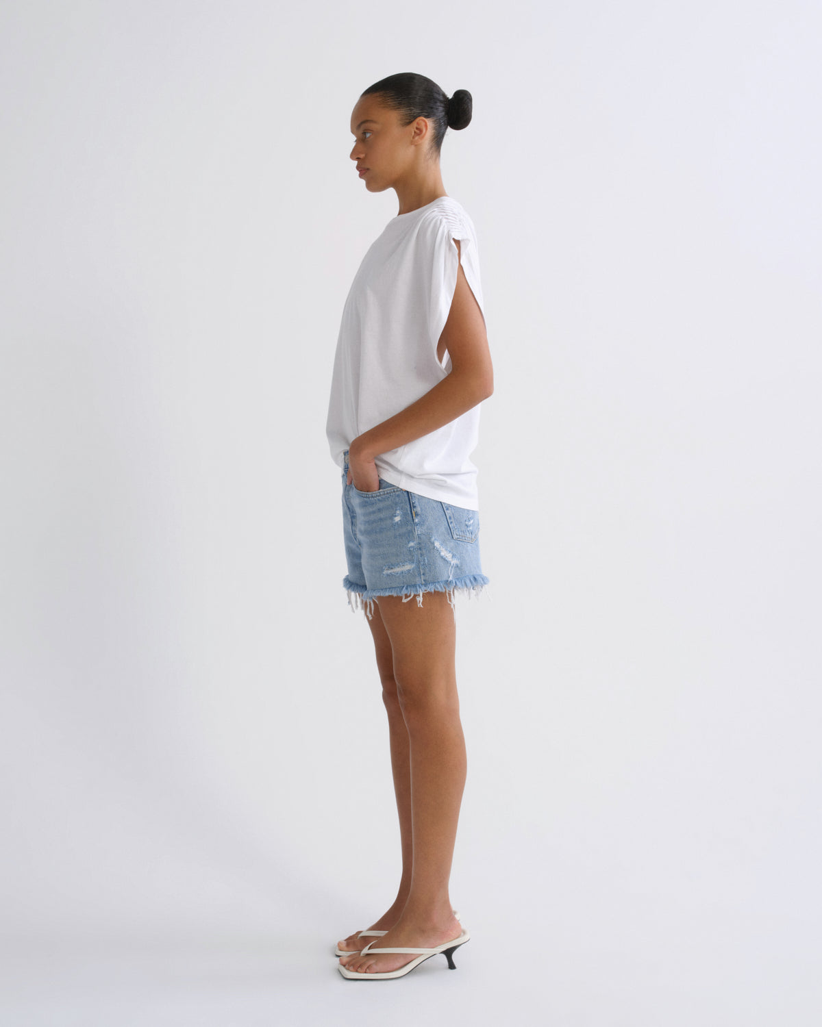 A model shown in profile wearing a white oversized sleeveless top, light-wash distressed denim shorts, and clear-strap heeled sandals against a plain light gray studio background.