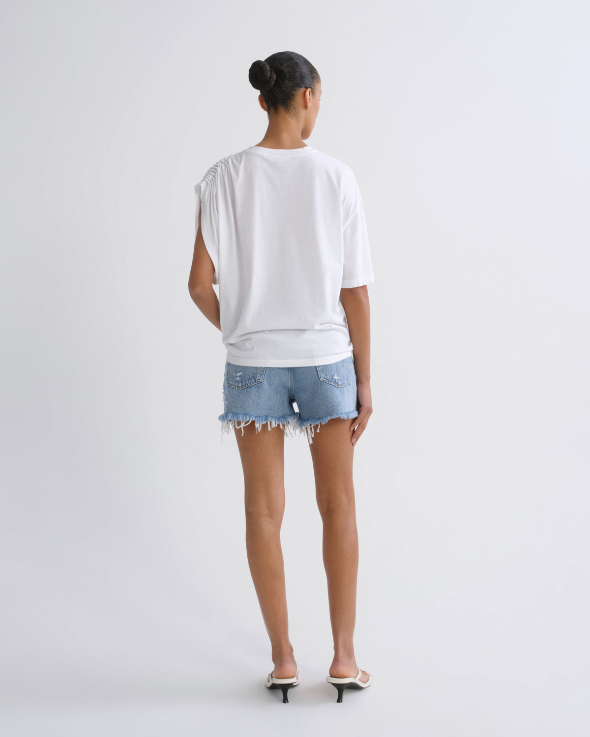 A woman viewed from behind wearing a white oversized short-sleeve top, light blue denim shorts with frayed hems, and white heeled sandals against a plain light gray studio background.