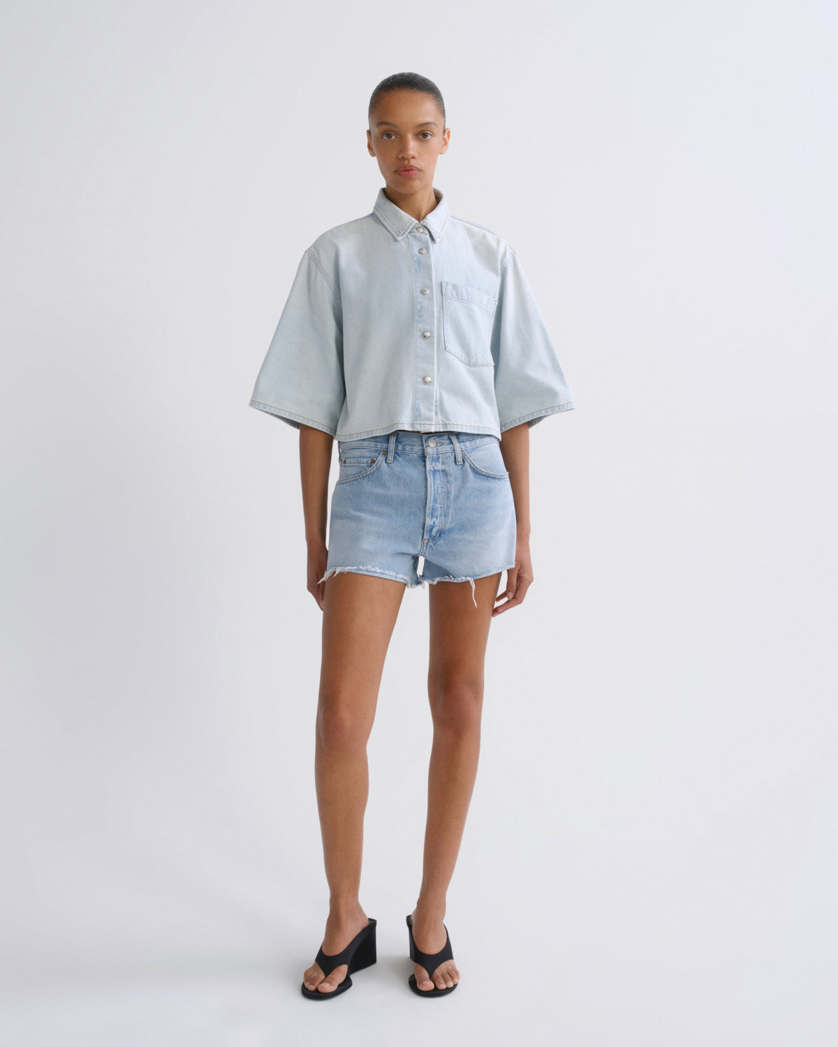 A model stands against a light gray studio background wearing a light blue cropped short-sleeve button-up shirt, light-wash denim shorts, and black sandals.