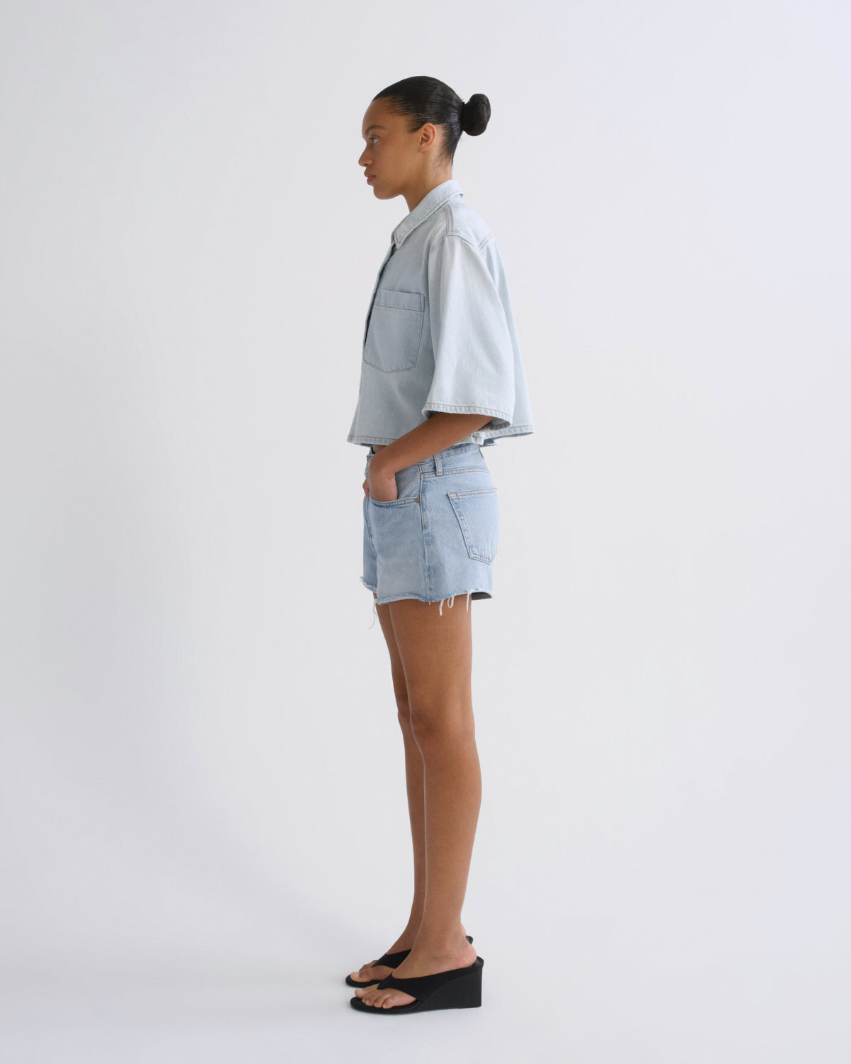 A model shown in profile wearing a light blue cropped denim shirt and matching light blue denim shorts, paired with black wedge sandals, against a plain white studio background.