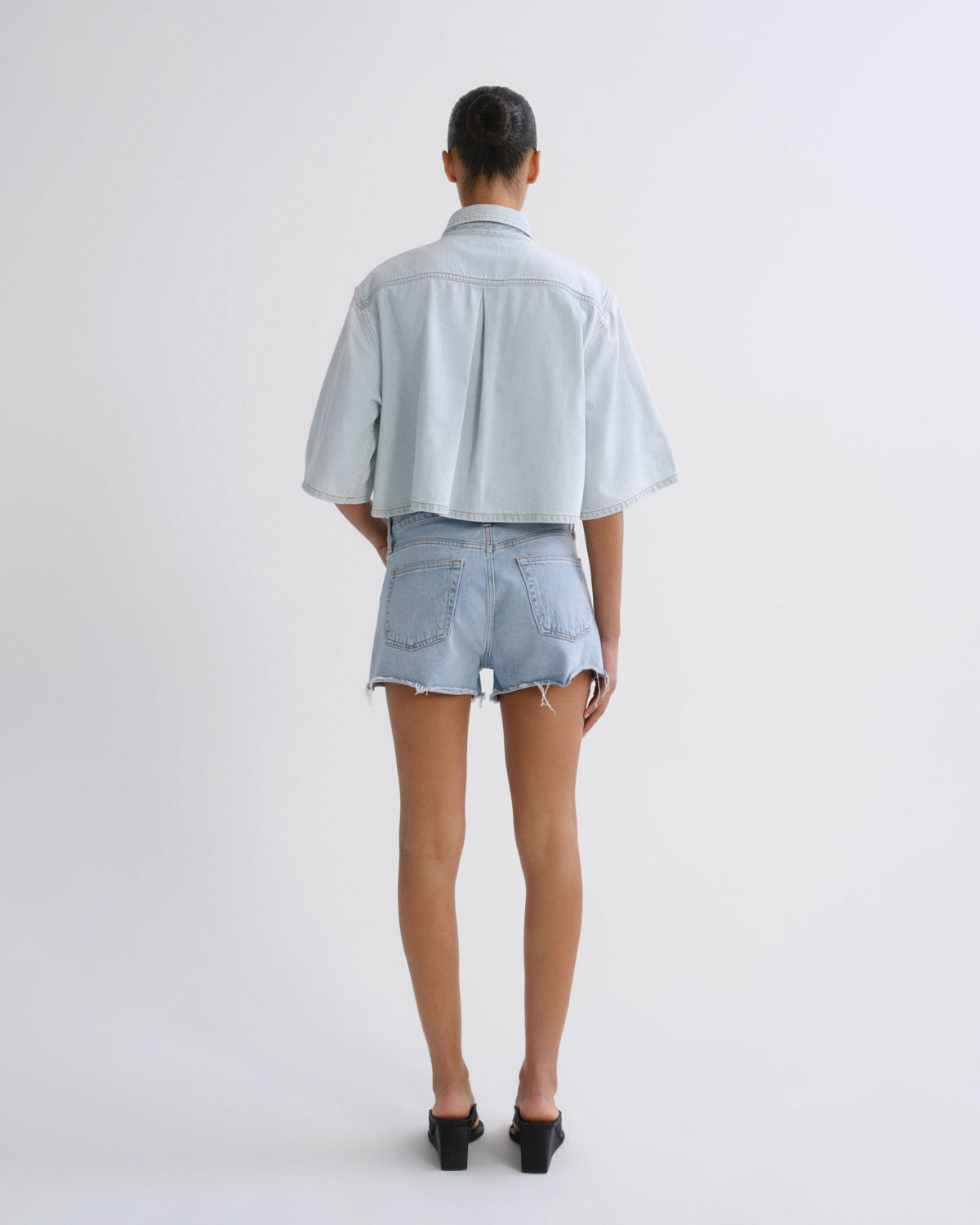A model viewed from the back wearing a light blue cropped denim shirt and matching light blue denim shorts, paired with black mule heels, against a plain light gray studio background.