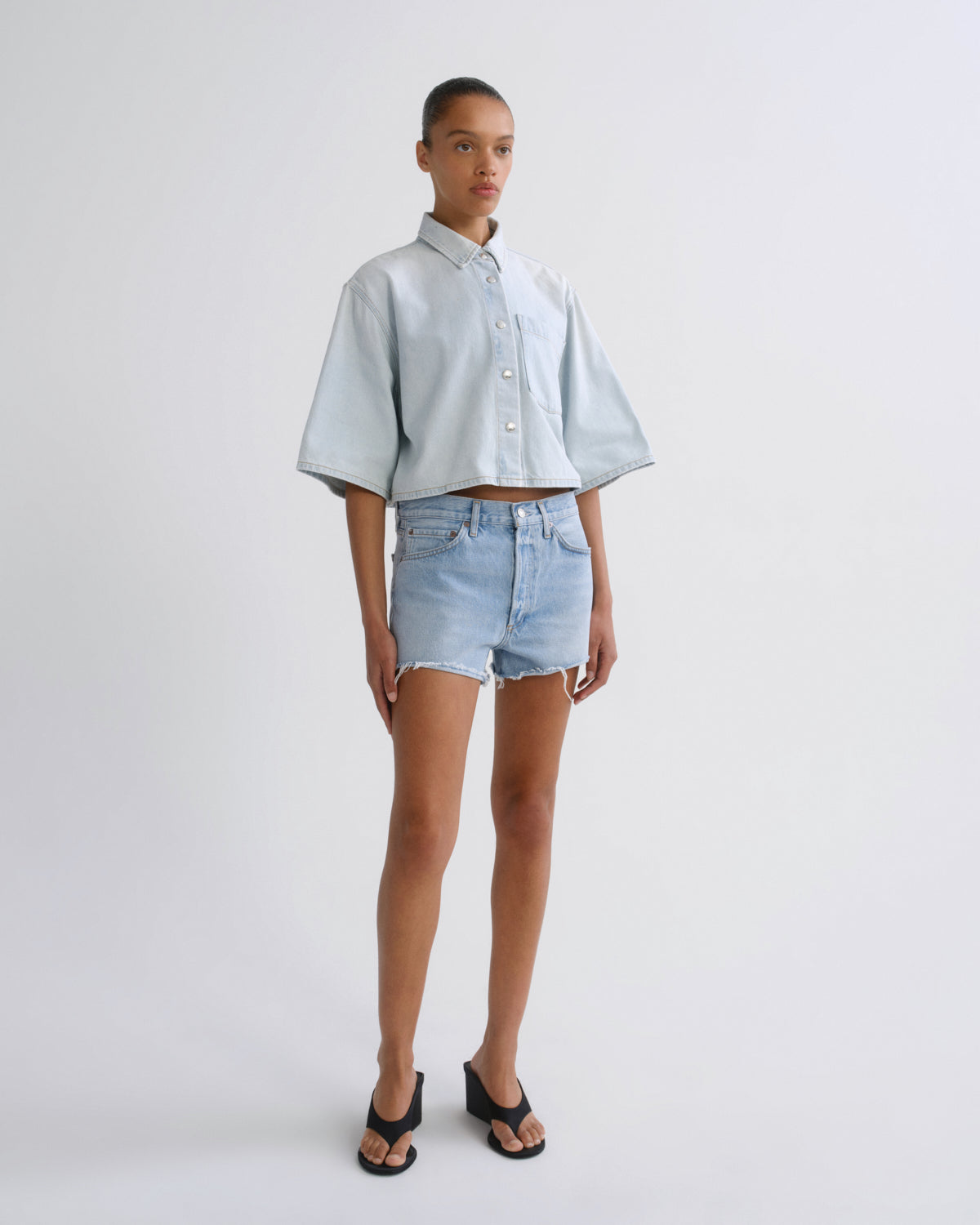 A model stands against a light gray studio background wearing a light blue cropped button-up shirt with wide short sleeves, light-wash denim shorts, and black slide sandals.