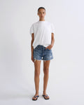A model stands against a light gray studio background wearing a white short-sleeve t-shirt, blue denim shorts with frayed hems, and blue flip-flop sandals.