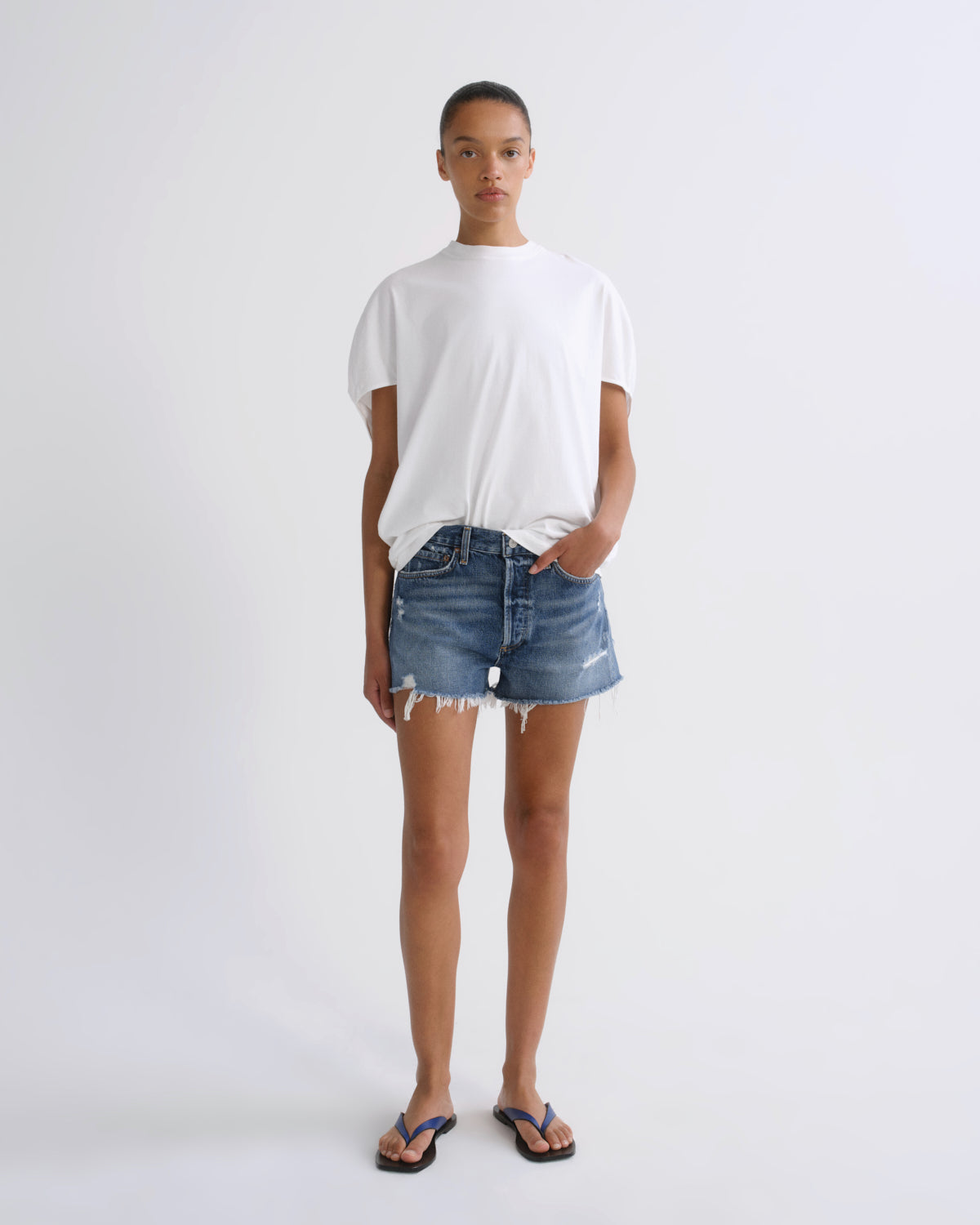 A model stands against a light gray studio background wearing a white short-sleeve t-shirt, blue denim shorts with frayed hems, and blue flip-flop sandals.