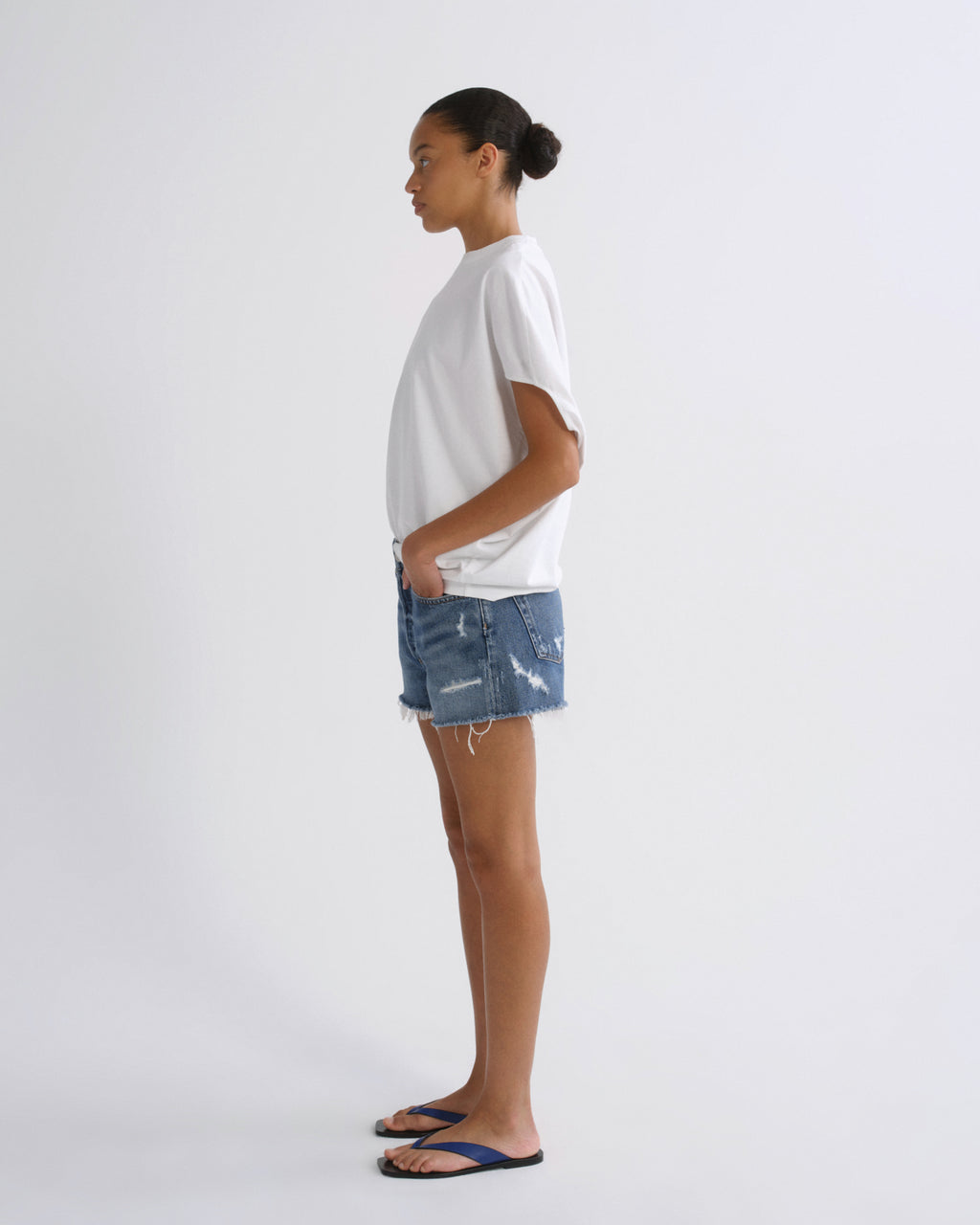 A model standing in profile wearing a white t-shirt, distressed blue denim shorts, and dark blue flip-flops against a plain light gray studio background.