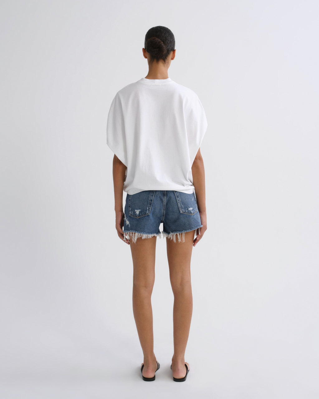 A model viewed from behind wearing a white oversized short-sleeve t-shirt and distressed denim shorts with frayed hems, standing against a light gray studio background.