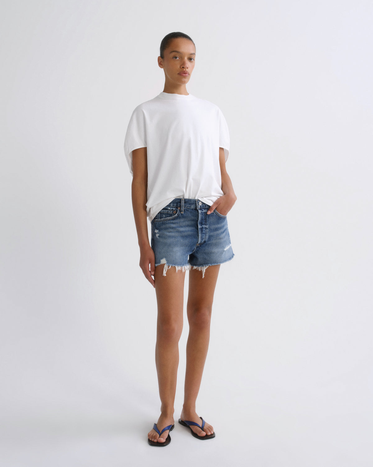 A model stands against a light gray studio background wearing a white short-sleeve t-shirt, distressed denim shorts, and blue flip-flops.