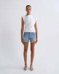 A model stands against a light gray studio background wearing a white asymmetrical sleeveless top, light-wash distressed denim shorts, and white strappy high-heel sandals.