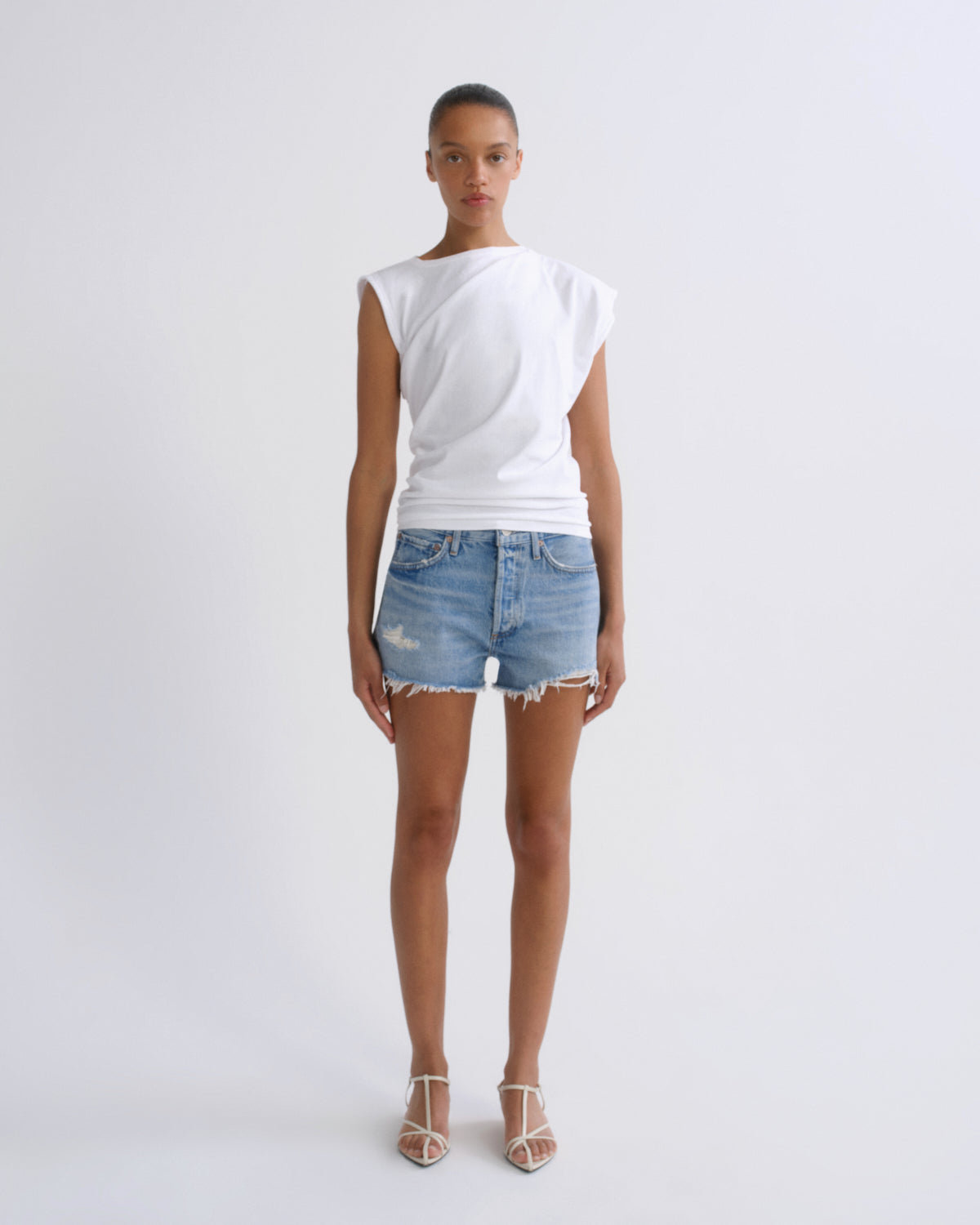 A model stands against a light gray studio background wearing a white asymmetrical sleeveless top, light-wash distressed denim shorts, and white strappy high-heel sandals.