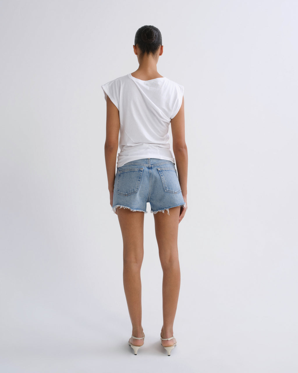 A model viewed from behind wearing a white draped sleeveless top, light-wash denim shorts, and white heels against a plain light gray studio background.