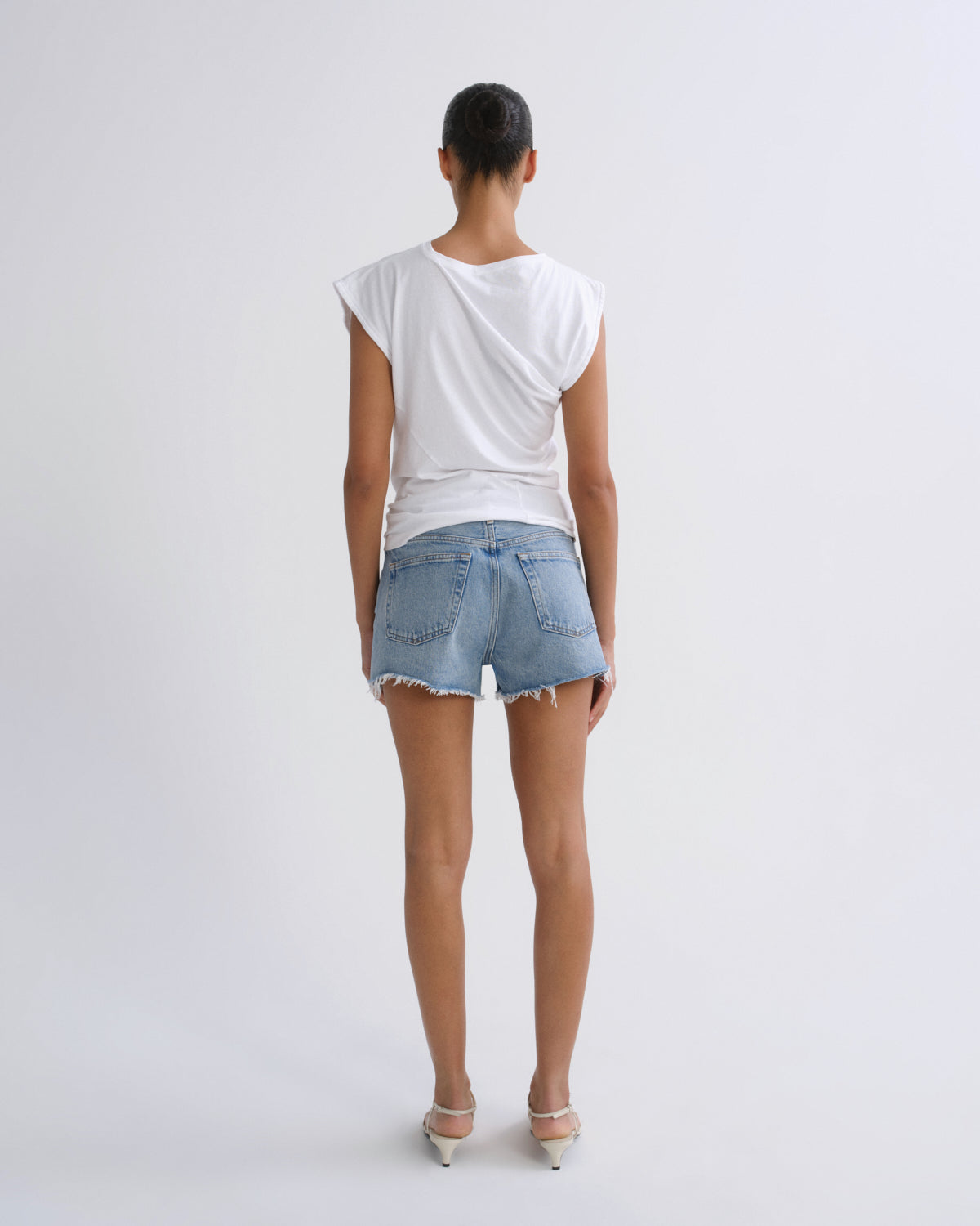 A model viewed from behind wearing a white draped sleeveless top, light-wash denim shorts, and white heels against a plain light gray studio background.