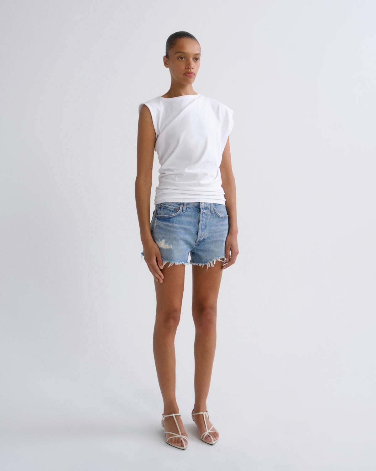 A model stands against a light gray studio background wearing a white sleeveless wrap-style top, light blue distressed denim shorts, and clear-strap heeled sandals.