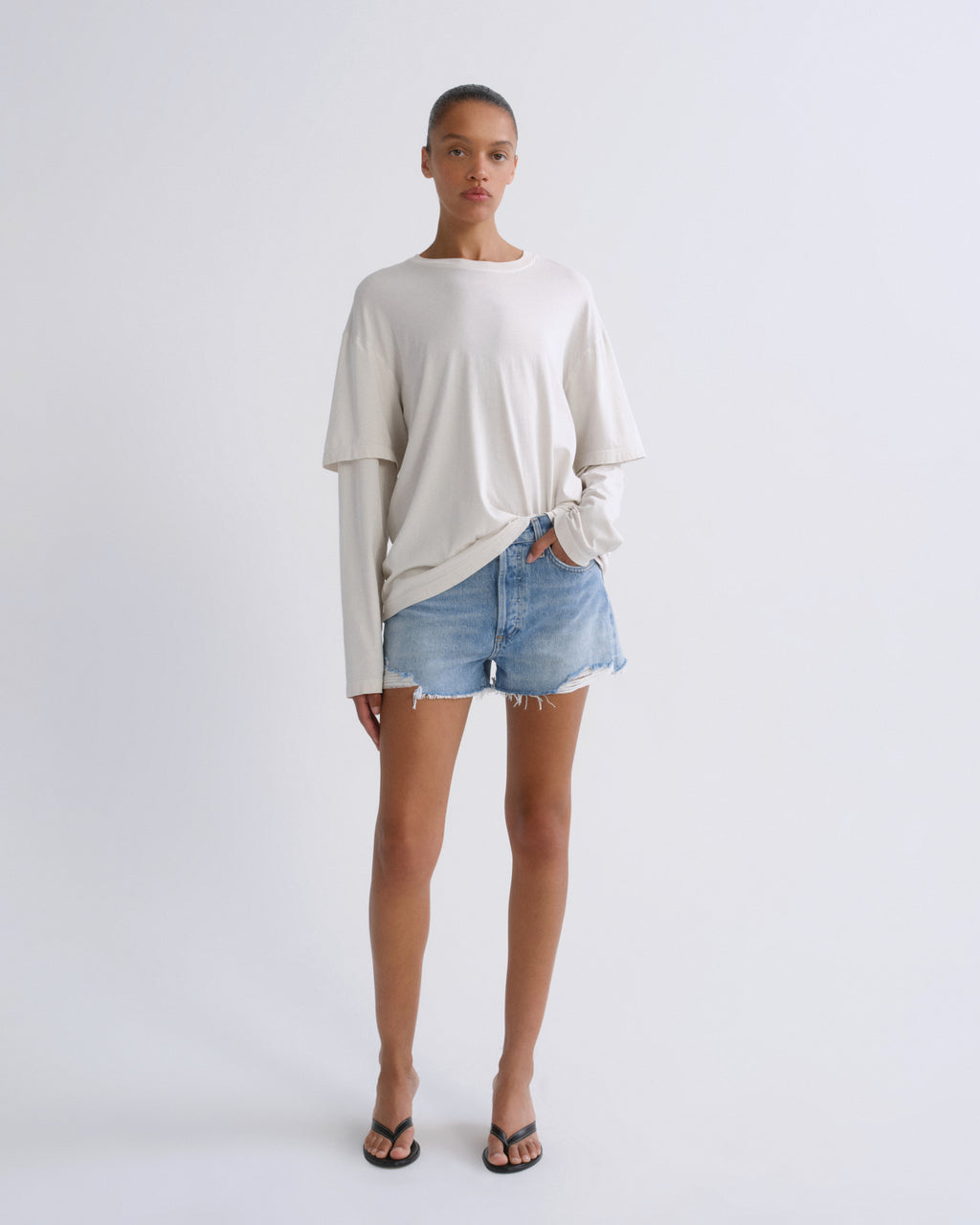 A model stands against a light gray studio background wearing a cream-colored long-sleeve t-shirt with a twisted hem, light-wash denim shorts, and black flip-flop sandals.