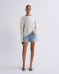 A model stands against a light gray studio background wearing a cream-colored long-sleeve t-shirt with a twisted hem, light-wash denim shorts, and black flip-flop sandals.