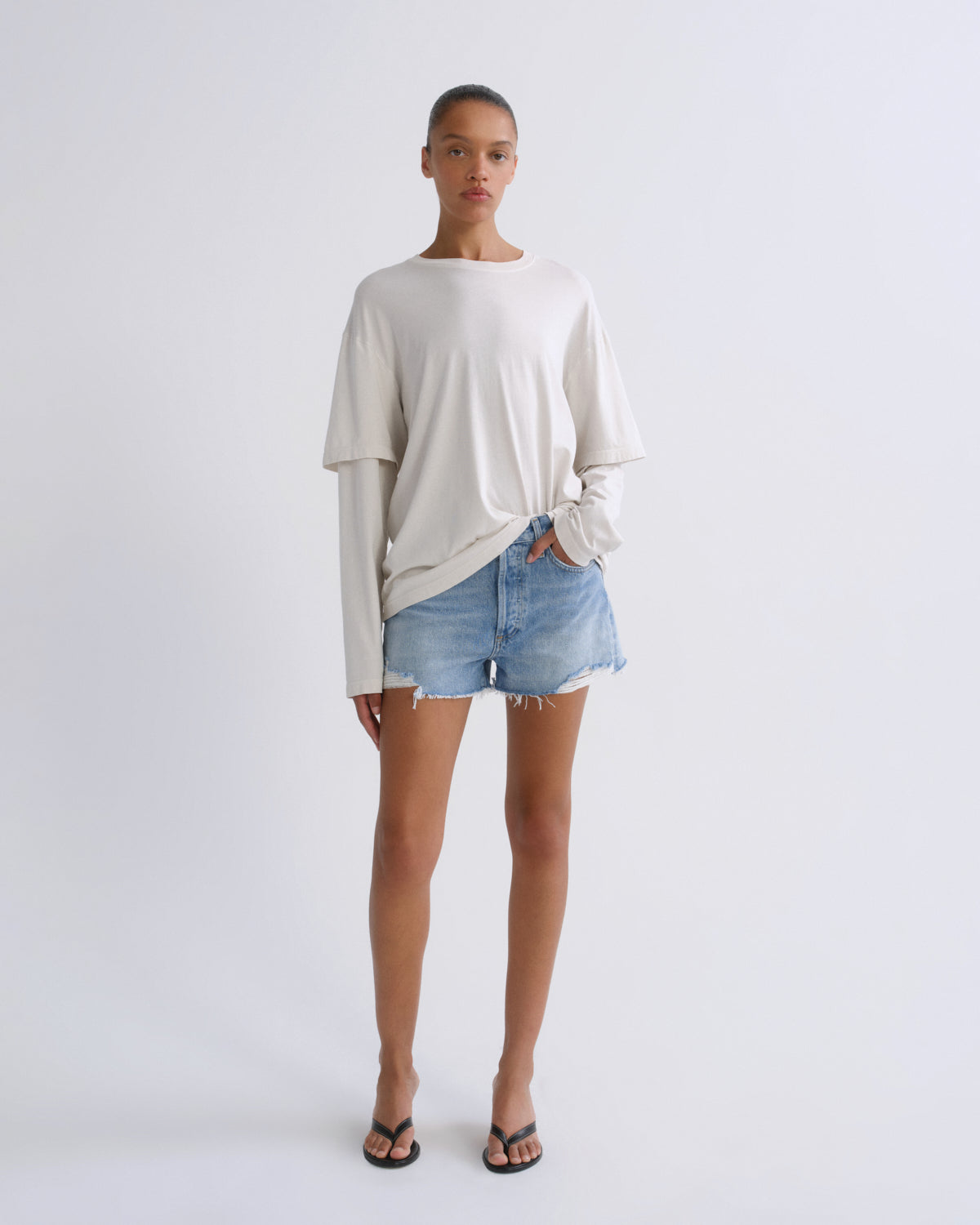 A model stands against a light gray studio background wearing a cream-colored long-sleeve t-shirt with a twisted hem, light-wash denim shorts, and black flip-flop sandals.
