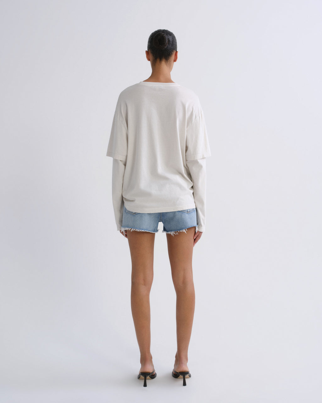 A model viewed from behind wearing an oversized off-white long-sleeve t-shirt, light-wash denim shorts, and black high-heeled sandals against a plain light gray studio background.
