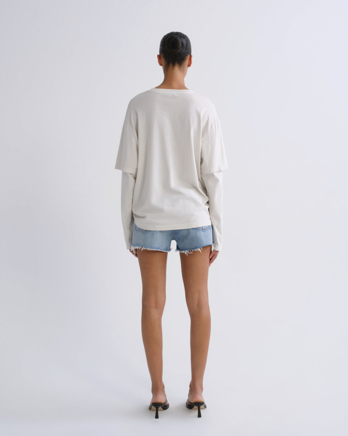 A model viewed from behind wearing an oversized off-white long-sleeve t-shirt, light-wash denim shorts, and black high-heeled sandals against a plain light gray studio background.