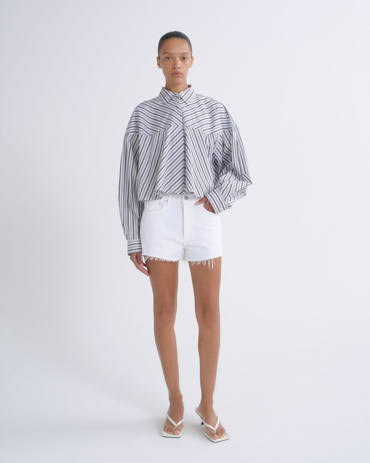 A model stands against a light gray studio background wearing a cropped, long-sleeve striped button-up shirt with white frayed denim shorts and white heeled sandals.