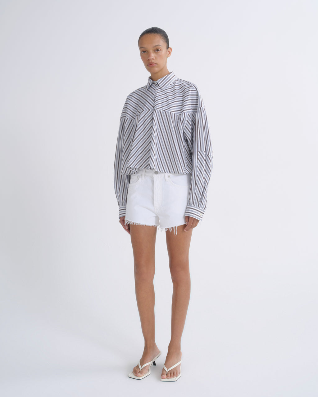 A model stands against a light gray studio background wearing a long-sleeve button-up shirt with vertical black and white stripes, white denim shorts, and white thong sandals.