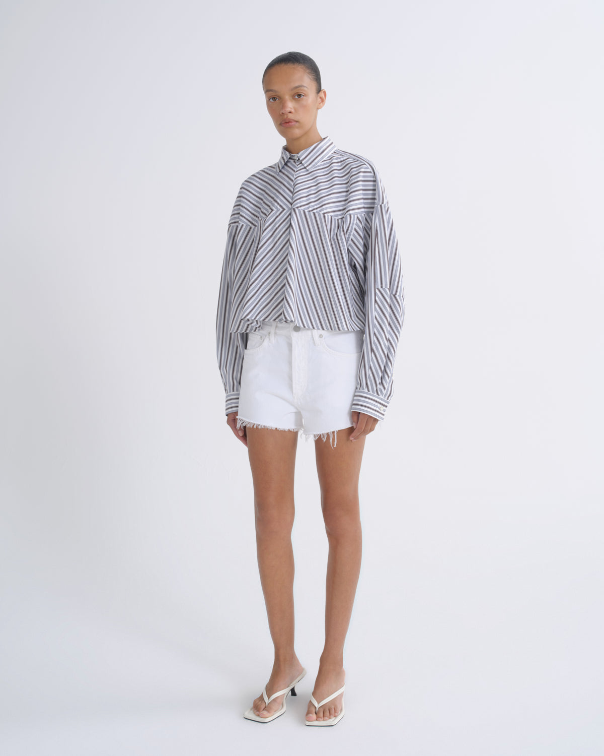 A model stands against a light gray studio background wearing a long-sleeve button-up shirt with vertical black and white stripes, white denim shorts, and white thong sandals.