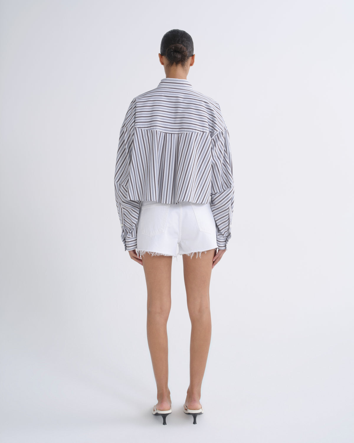 A model viewed from the back wearing a cropped, long-sleeve striped button-up shirt with white shorts and white pointed-toe heels, set against a plain light gray studio background.