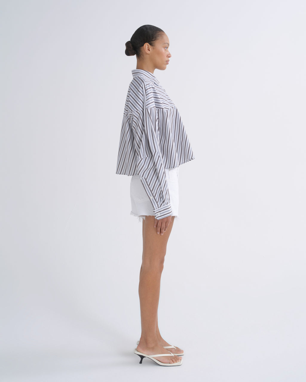 A woman stands in profile wearing a loose black-and-white vertically striped button-up shirt, white shorts, and clear slide sandals against a plain light gray studio background.