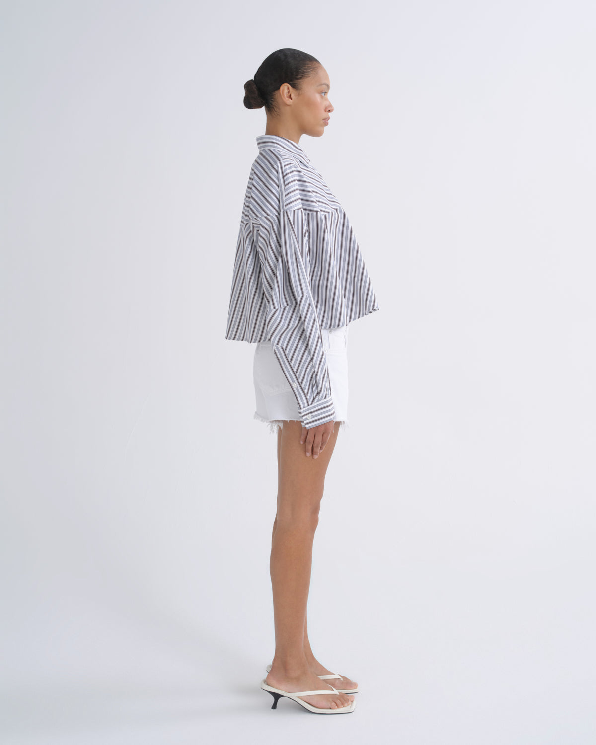 A woman stands in profile wearing a loose black-and-white vertically striped button-up shirt, white shorts, and clear slide sandals against a plain light gray studio background.