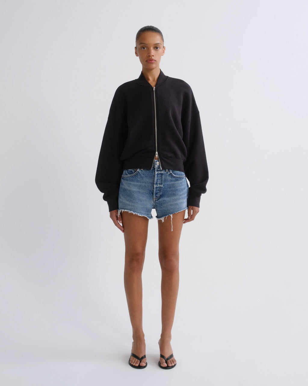 A model stands against a light gray studio background wearing a black zip-up bomber-style sweatshirt, blue denim shorts, and black sandals.